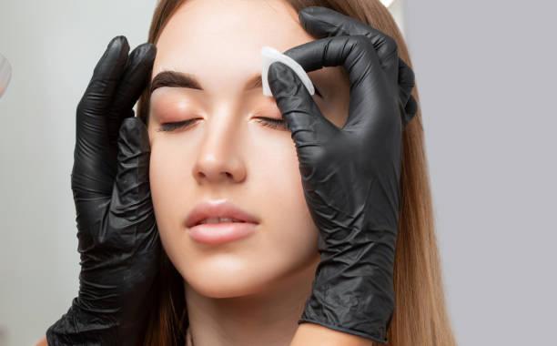 a person with black gloves wiping a woman's eyebrow with a cotton pad